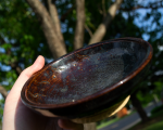 A hand holds a ceramic bowl in the sunshine revealing the blues and purples in a dark brownish glaze. Round divots decorate the rim of the bowl.